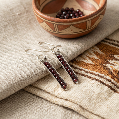 Silver earrings with red stones on a textured fabric surface with a bowl of beads in the background.
